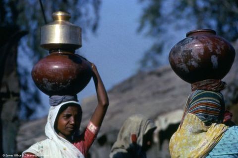 Two people wearing scarves covering their heads face away from us, balancing vessels on their heads. The person to our left carries what looks like a clay vessel on a head pad. The other reaches a dark-skinned hand up to balance a gold-coloured vessel with a round bottom, straight sides, and flaring mouth.